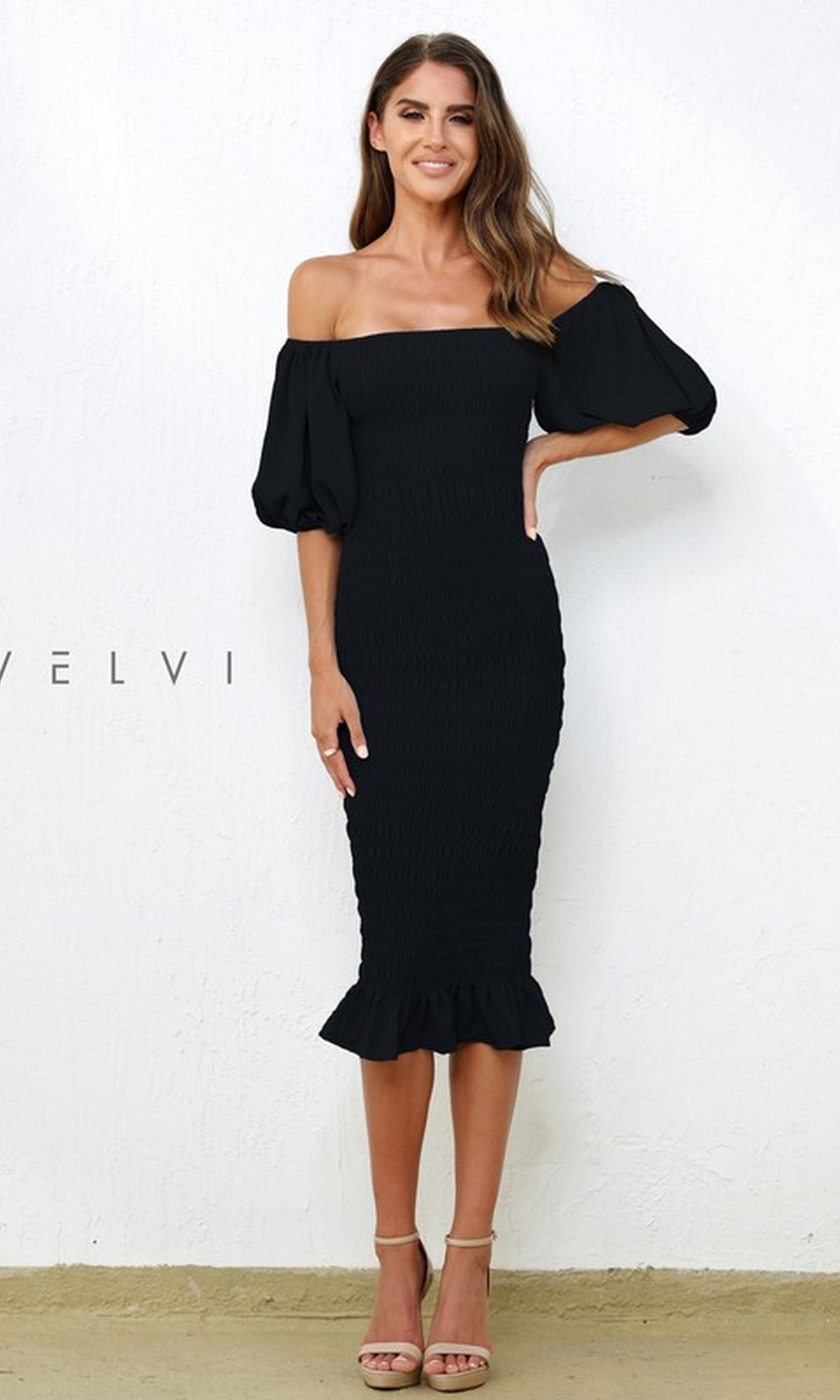 A woman stands against a white wall wearing the Skyla Knee-Length Puff-Sleeve Wedding-Guest Dress, featuring an off-the-shoulder neckline, puffed sleeves, ruffled hem, and paired with beige heeled sandals.